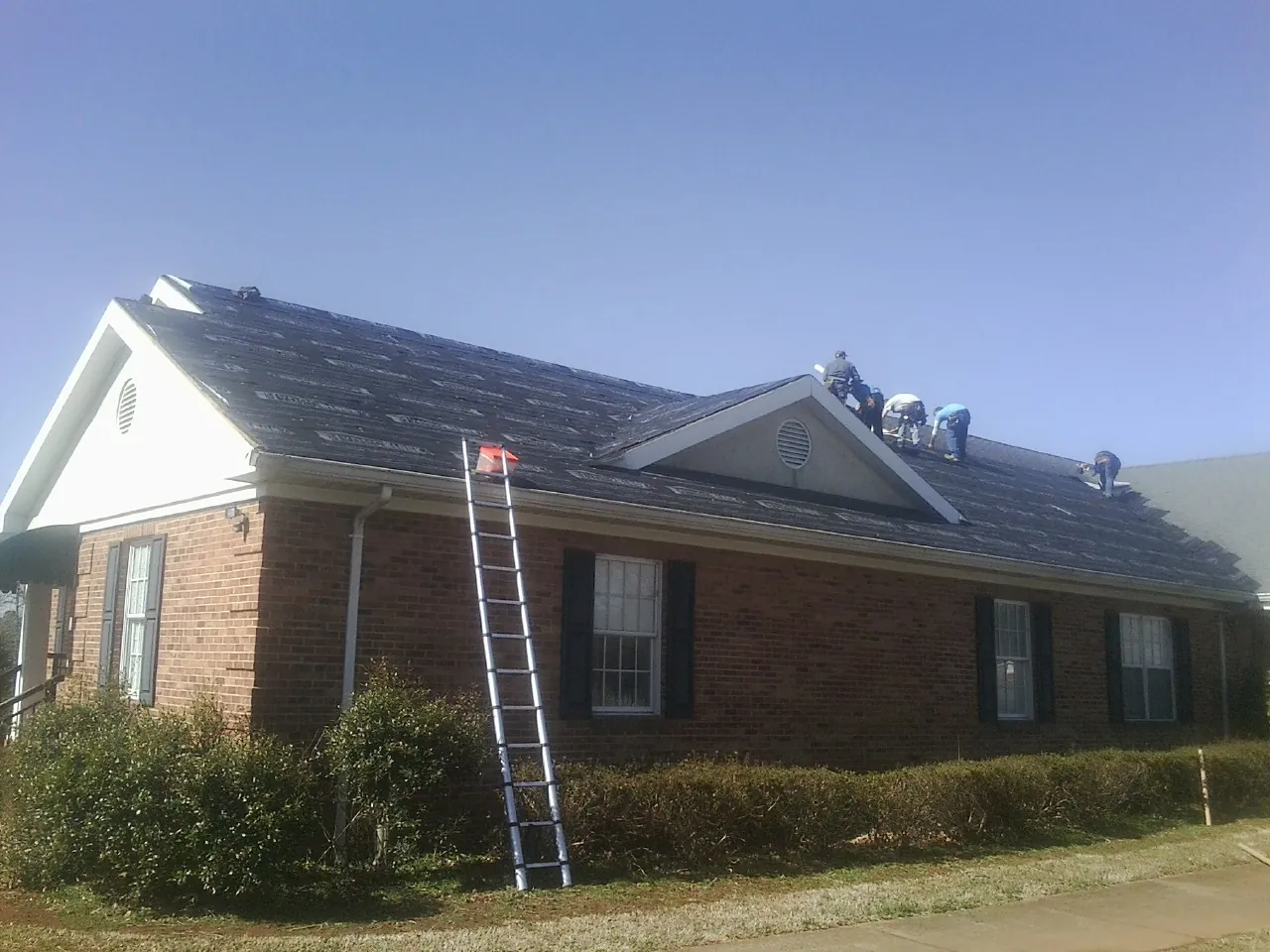 Crew tearing shingles off a brick ranch roof with a ladder set — Emergency Roof Repair work by our crew in Tougaloo