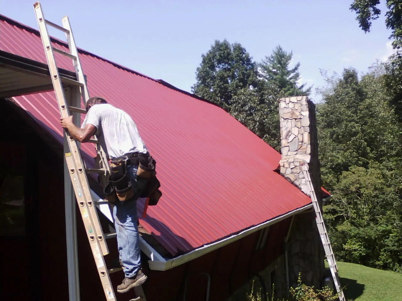 Roofer on a ladder beside a red metal roof and stone chimney — Soffit & Fascia Repair by Roofing Brothers in Tougaloo